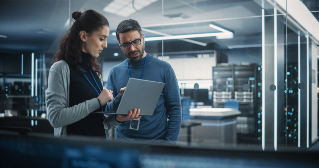 A man and woman in a server room look at a laptop together